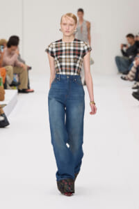 Model walks a white runway in a plaid short-sleeve top tucked into high-waisted wide-leg blue jeans, with floral platform shoes and gold bracelets.