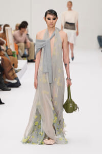 Model walks the runway in a sheer gray floral gown with a draped knit scarf, wearing small sunglasses and carrying a green handbag; audience visible in the background.