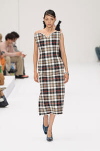 Model walks a runway wearing a plaid off-the-shoulder midi dress with blue heels, audience in background.**