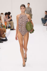 Model walks the runway in a taupe crocheted halter bodysuit, holding a green handbag under her arm.