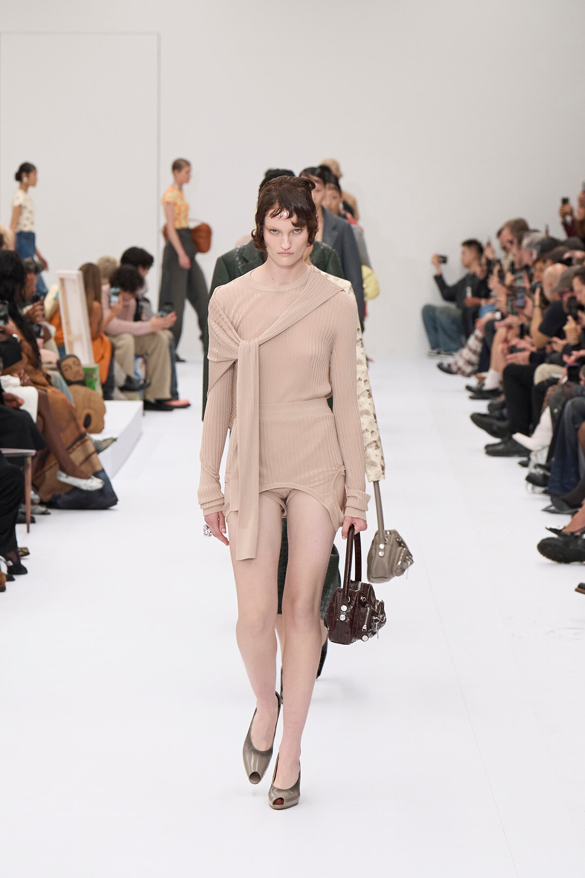 Model in a nude-toned knit outfit walking the runway, scarf draped across chest and beige heels, audience on both sides.