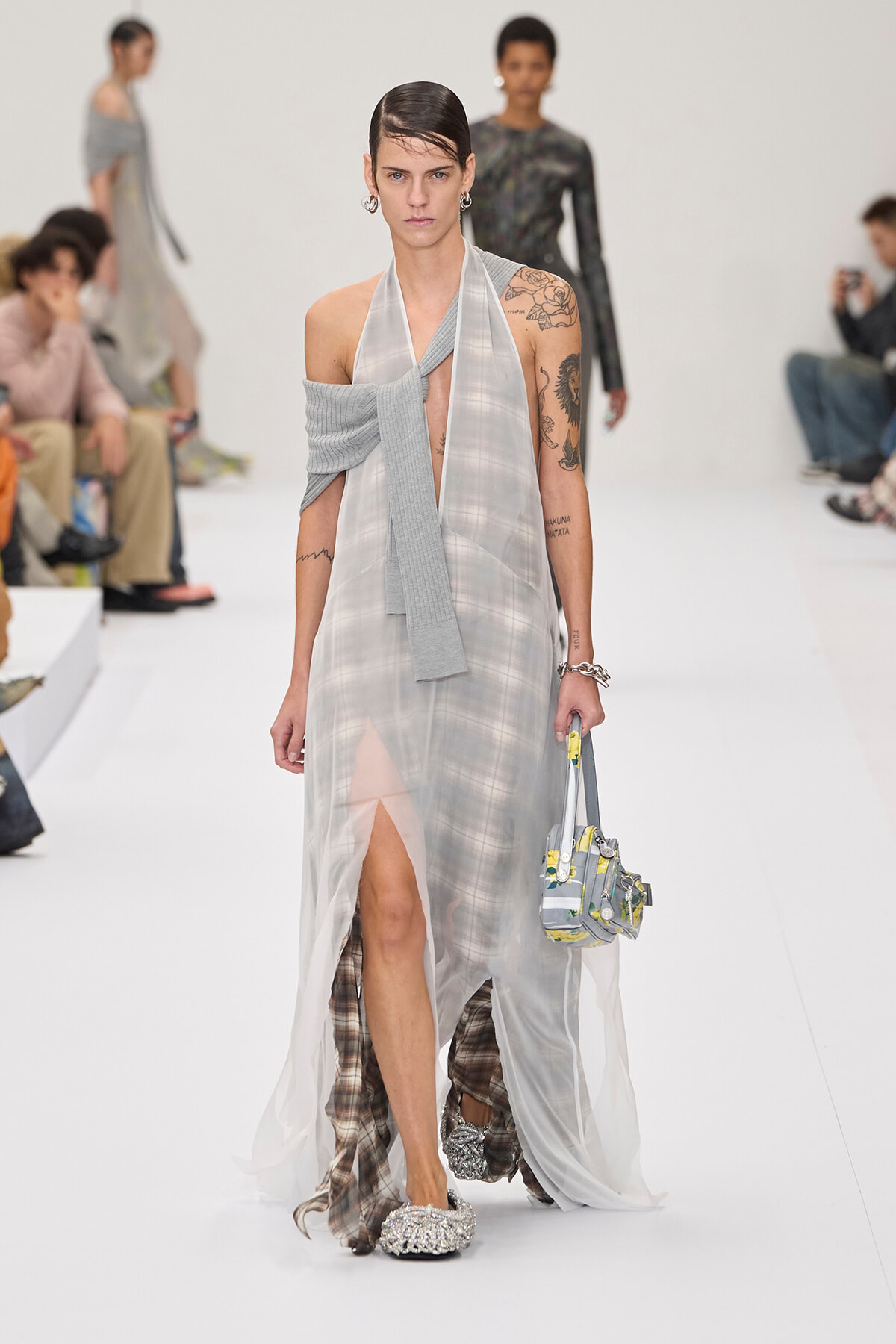Model walking a fashion runway in a sheer light-gray gown with a plaid underlay, a gray scarf draped across the shoulders, and a small patterned handbag.