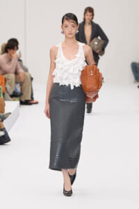 Model walking a runway in a white ruffled sleeveless top, dark crocodile-print skirt, black patent heels, and a tan handbag.