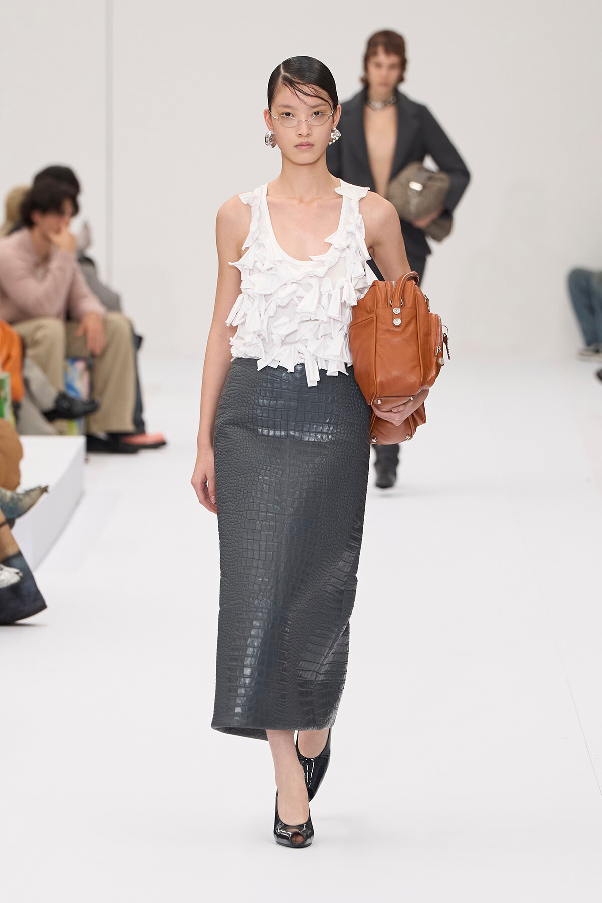 Model walking a runway in a white ruffled sleeveless top, dark crocodile-print skirt, black patent heels, and a tan handbag.