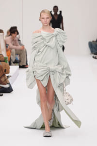 Model walks the runway in a pale mint off-shoulder gown with oversized front and lower bows, carrying a studded white bag.