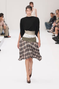 Model on a white runway wearing a black draped top with a multi-pattern wrap skirt and wide beige snakeskin belt, brown heels visible.