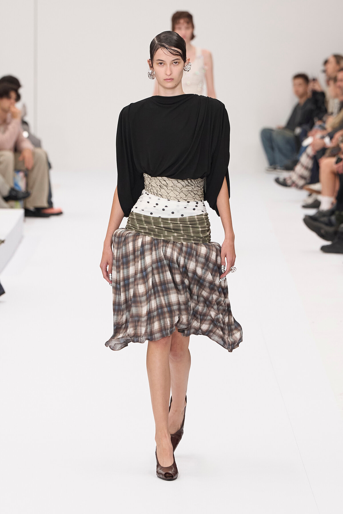 Model on a white runway wearing a black draped top with a multi-pattern wrap skirt and wide beige snakeskin belt, brown heels visible.