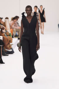 Model walking a white runway in a sleek black sleeveless jumpsuit with a V neck and sculpted waist; audience in the background.