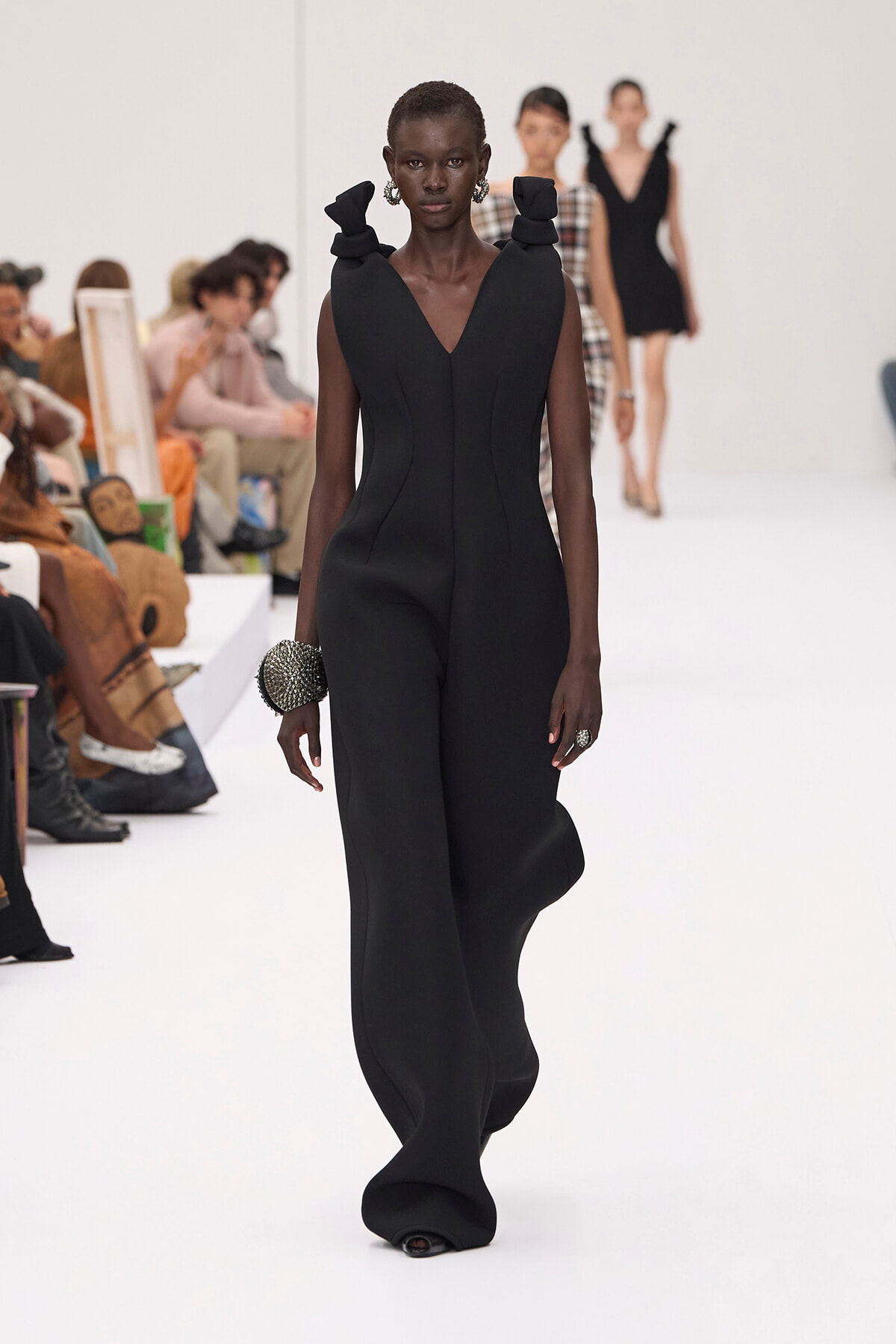 Model walking a white runway in a sleek black sleeveless jumpsuit with a V neck and sculpted waist; audience in the background.