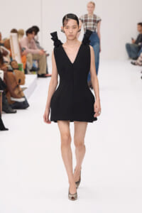 Model walks the runway in a sleek black V-neck mini dress with sculpted side seams and folded shoulder accents, neutral heels.