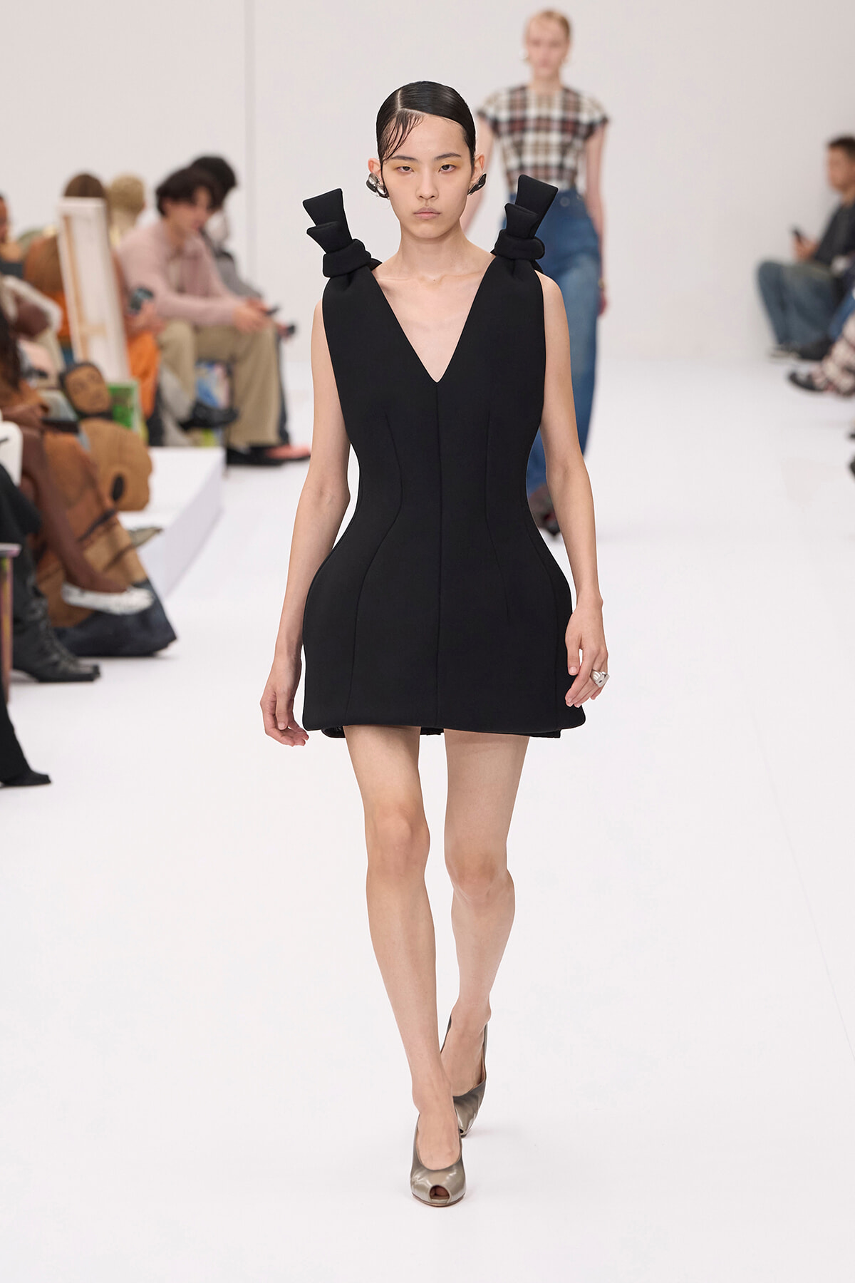 Model walks the runway in a sleek black V-neck mini dress with sculpted side seams and folded shoulder accents, neutral heels.