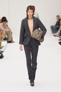 Model walks the runway in a dark gray oversized suit with the blazer open, carrying a taupe quilted handbag and wearing a chunky black-silver necklace.
