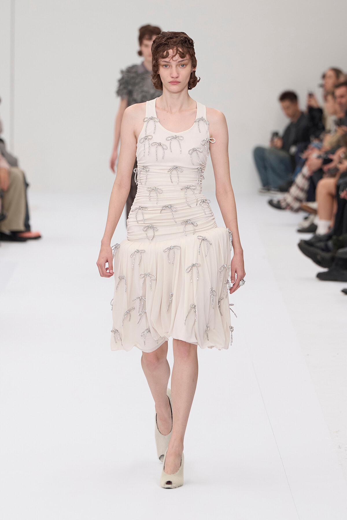 Fashion model walks the runway in an ivory sleeveless dress adorned with silver bow appliqués and matching heels.