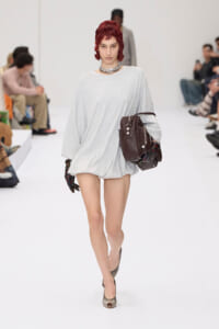 Model walking a white runway in a loose light-gray sweater dress, chunky silver necklace, red-streaked hair, carrying a brown leather backpack and a scarf, wearing taupe heels.