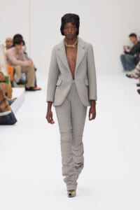 Model on a white runway wearing a light gray suit with an open front, gold chunky necklace, and ruched pants.