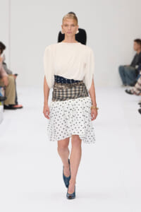Model walking a fashion runway in a cream draped top, layered plaid and polka-dot skirt, blue heels, and bold earrings.