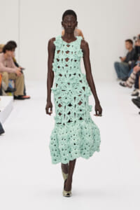 Model on a runway wearing a mint green crocheted sleeveless dress with a scalloped, floral pattern.