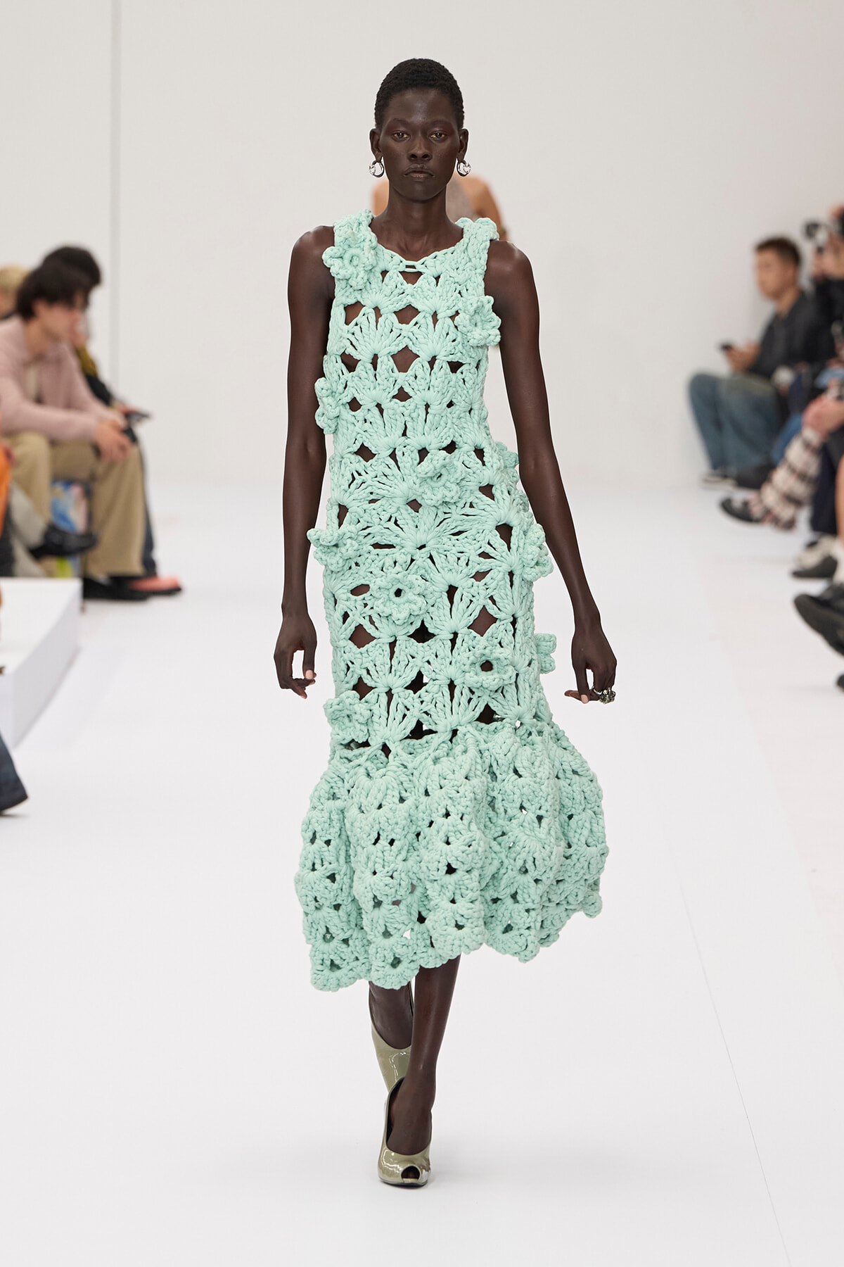 Model on a runway wearing a mint green crocheted sleeveless dress with a scalloped, floral pattern.