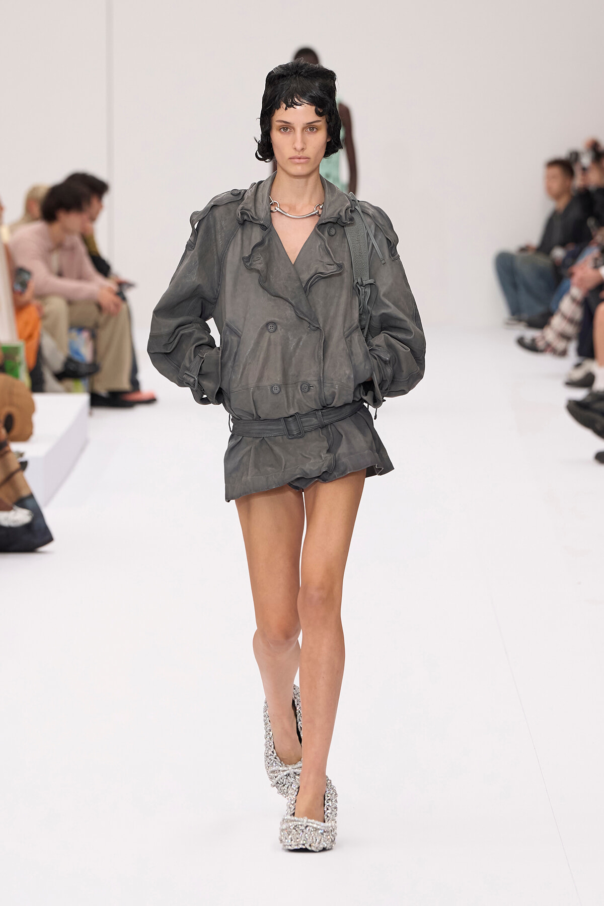 Model walking the runway in an oversized gray trench-style jacket with a belt and matching gray shorts; silver embellished heels visible on her feet.