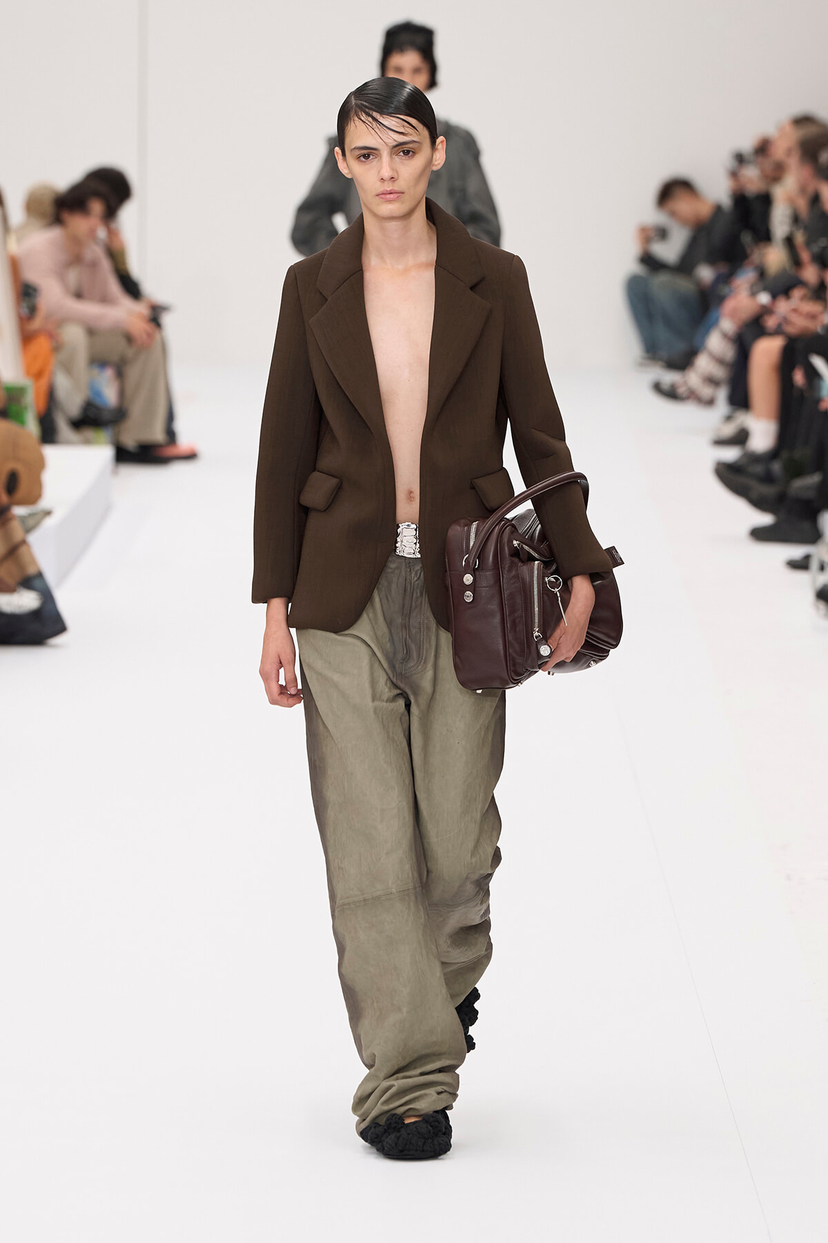 Model walking a white runway in a brown blazer, open at the chest, olive baggy pants, and a dark burgundy handbag tucked under her arm, audience watching.