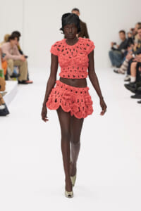 Black model walks the runway in coral pink crocheted two-piece: a cropped top and scalloped mini skirt, on a white catwalk with spectators