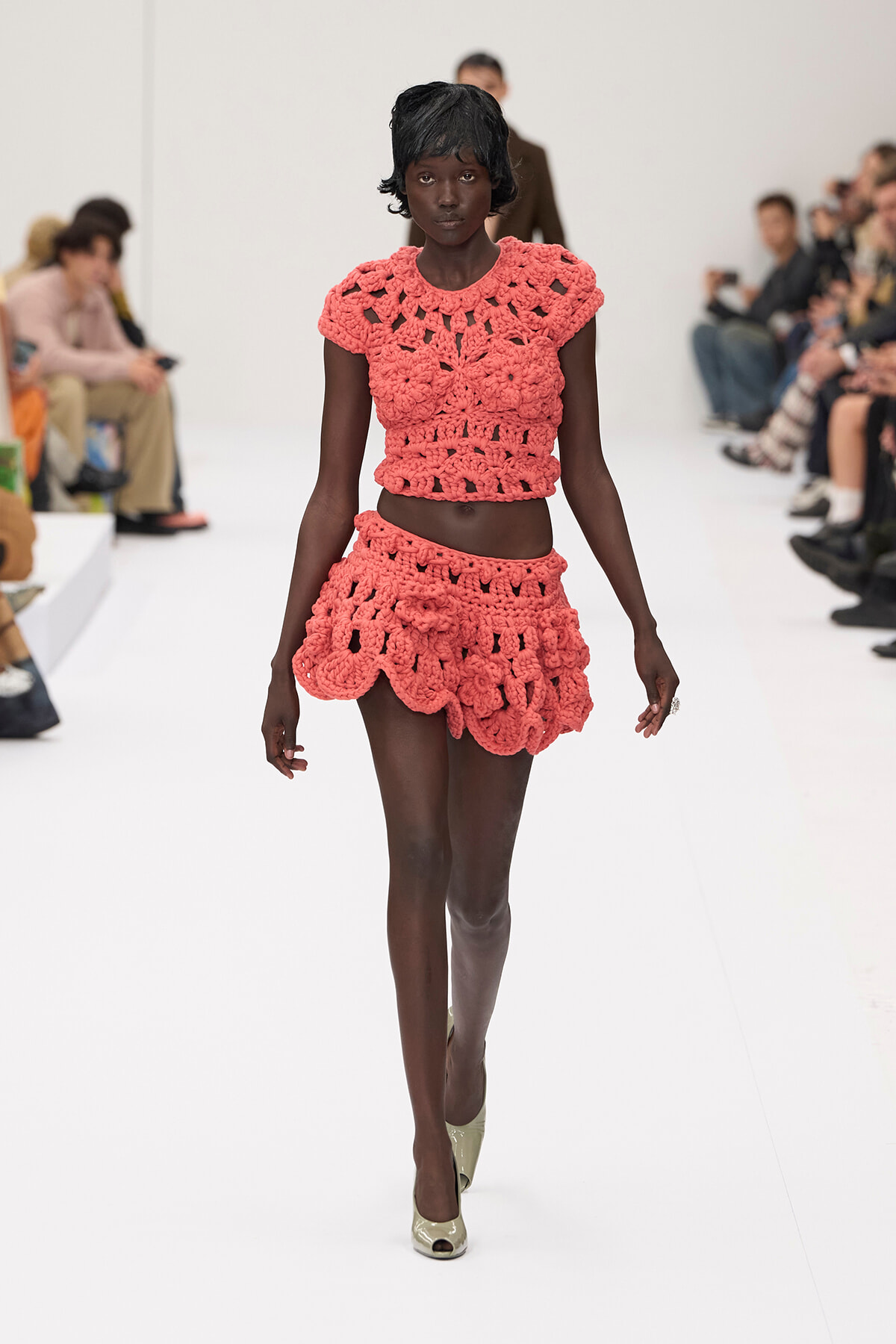 Black model walks the runway in coral pink crocheted two-piece: a cropped top and scalloped mini skirt, on a white catwalk with spectators
