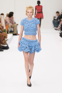 Model walks the runway in a light blue crocheted two-piece outfit featuring a cropped top and ruffled mini skirt; audience seated along the sides.