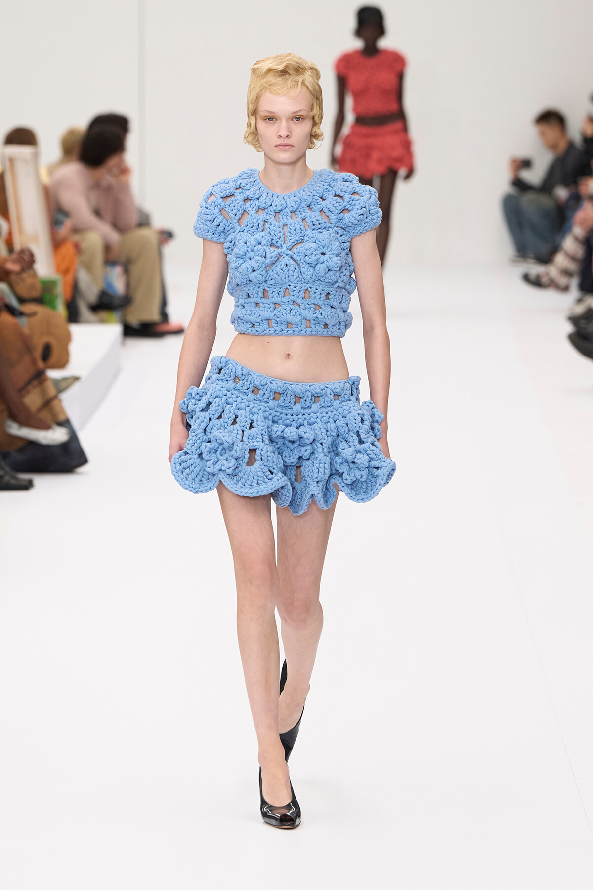 Model walks the runway in a light blue crocheted two-piece outfit featuring a cropped top and ruffled mini skirt; audience seated along the sides.