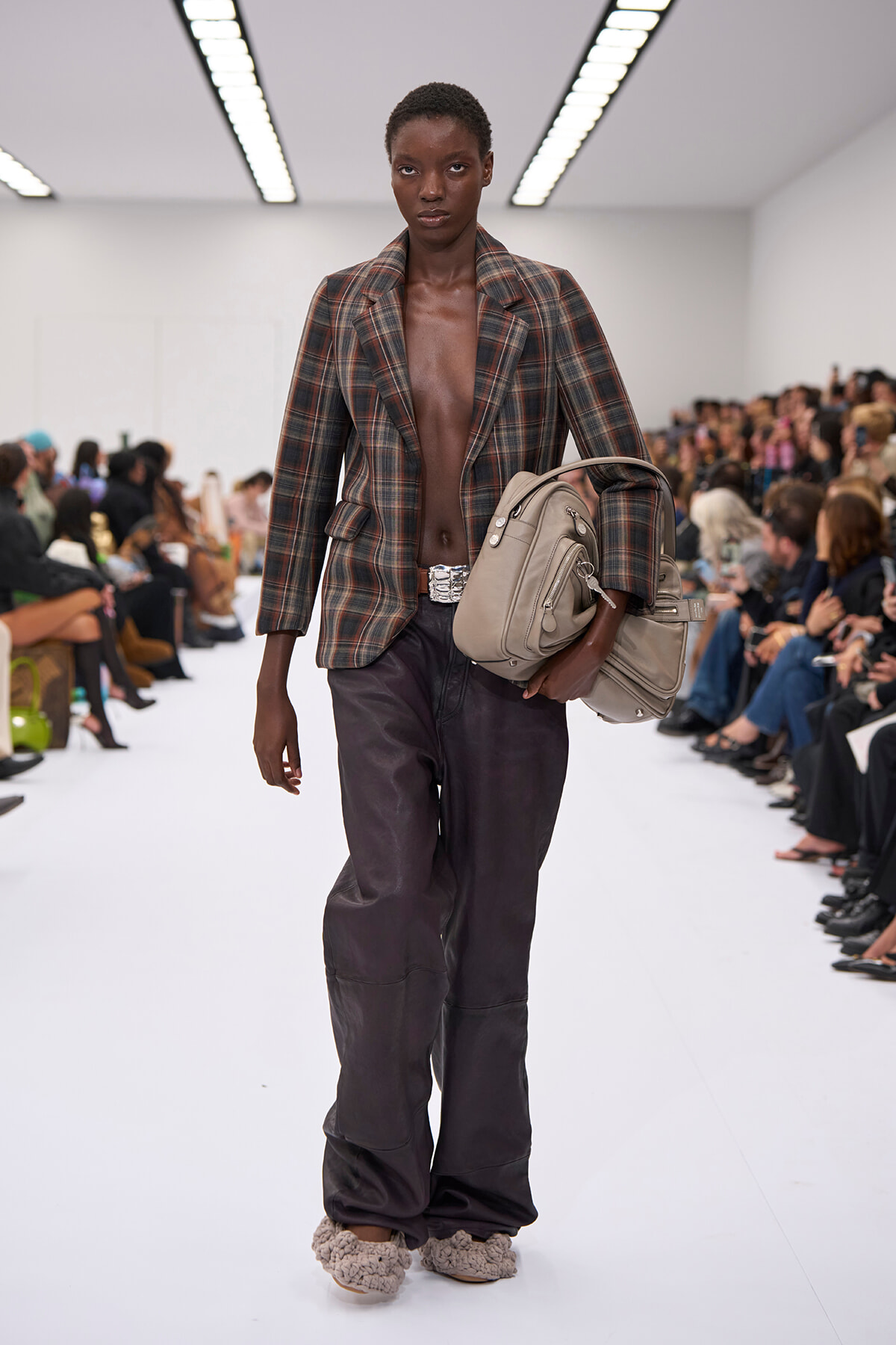 Runway model wearing an open plaid blazer with no top, dark wide-leg pants, a silver belt, beige handbag, and fuzzy slippers, with spectators in the background.