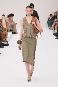 Runway model in an olive-green sleeveless vest and midi skirt, carrying a brown leather backpack on her shoulder.