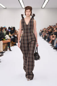 Model walking a fashion show in a brown plaid V-neck jumpsuit with bow-tied shoulders, carrying a small patterned handbag.