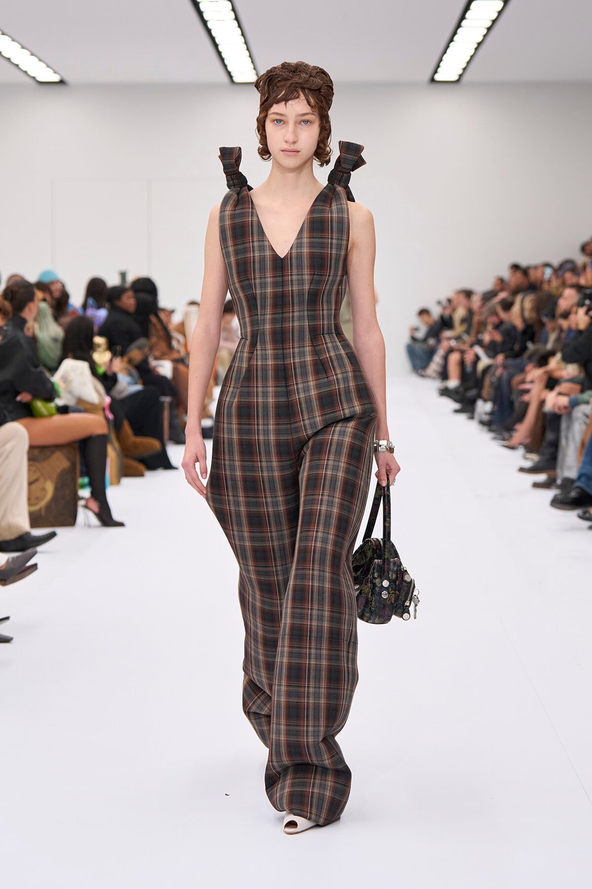 Model walking a fashion show in a brown plaid V-neck jumpsuit with bow-tied shoulders, carrying a small patterned handbag.