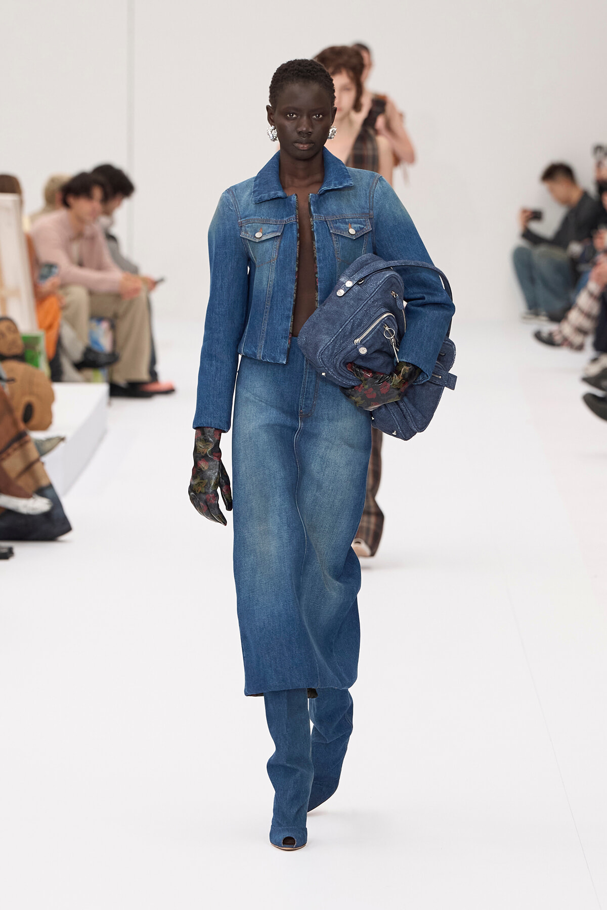 Black model walks the runway in a blue denim jacket and wide-leg jeans, carrying a stack of denim bags.