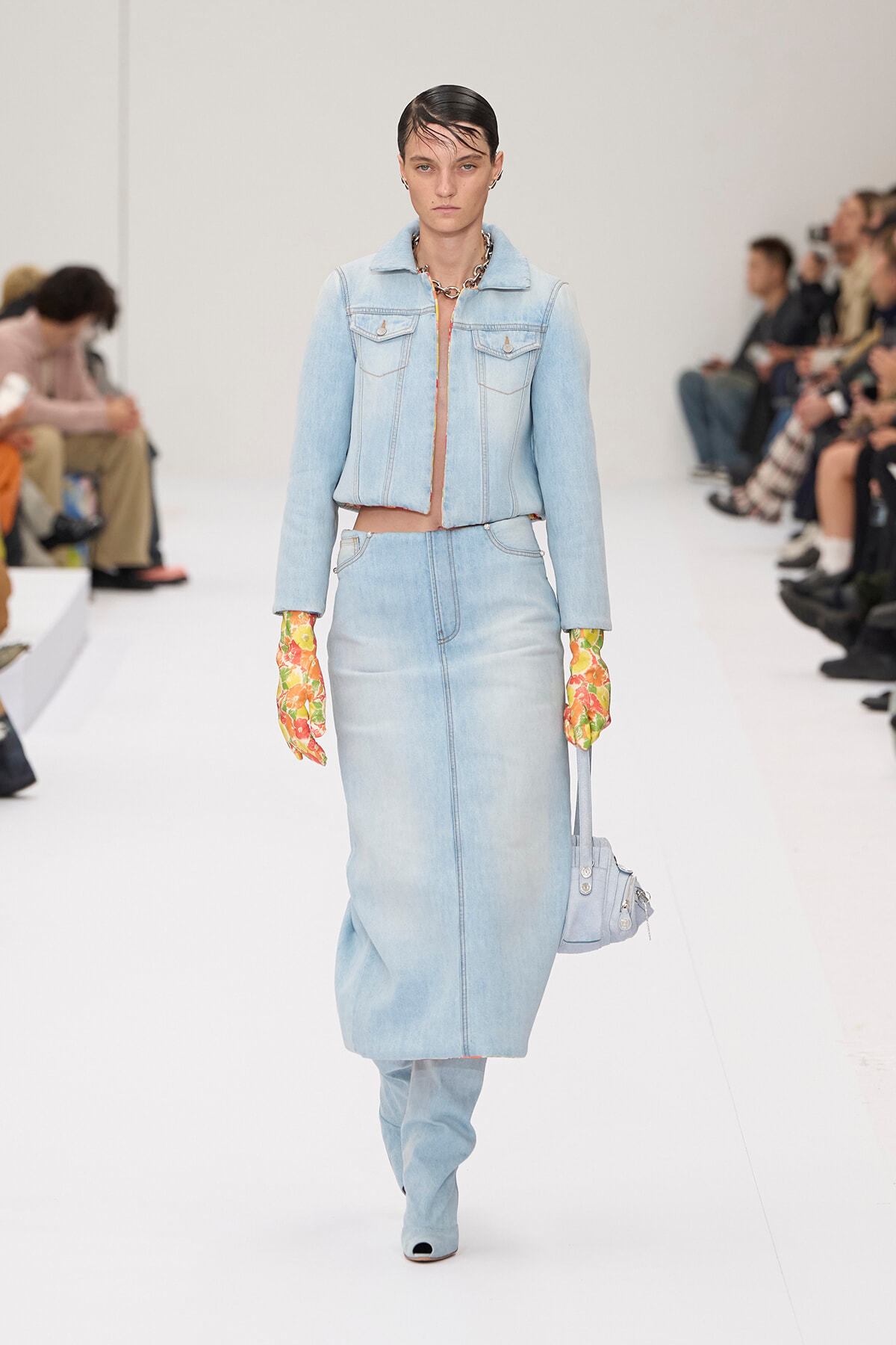 Model walking a runway in a light denim two-piece set with a cropped jacket and long skirt, accessorized by floral gloves and a chunky necklace.