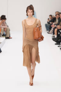 Model walks a fashion show in a beige sleeveless draped dress, carrying a brown leather handbag. Audience sits along a white runway.