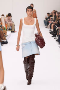 Model walking a fashion runway in a white scoop-neck tank, light denim skirt, tall brown knee-high boots, and a burgundy handbag. Silver earrings complete the look.