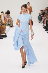 Model walking a runway in a pale blue, high-neck draped gown, holding extra fabric in one hand with a chunky silver bracelet visible on the wrist.