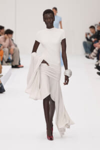 Black model walks the white runway in an ivory draped gown, holding fabric at the waist with a large crystal bracelet on her wrist.