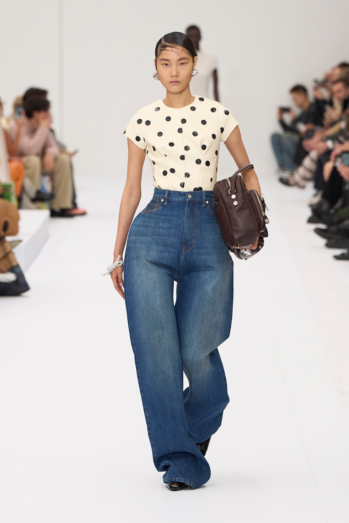 Model walks the runway in a cream polka-dot top and ultra-wide denim pants, carrying a dark brown handbag.