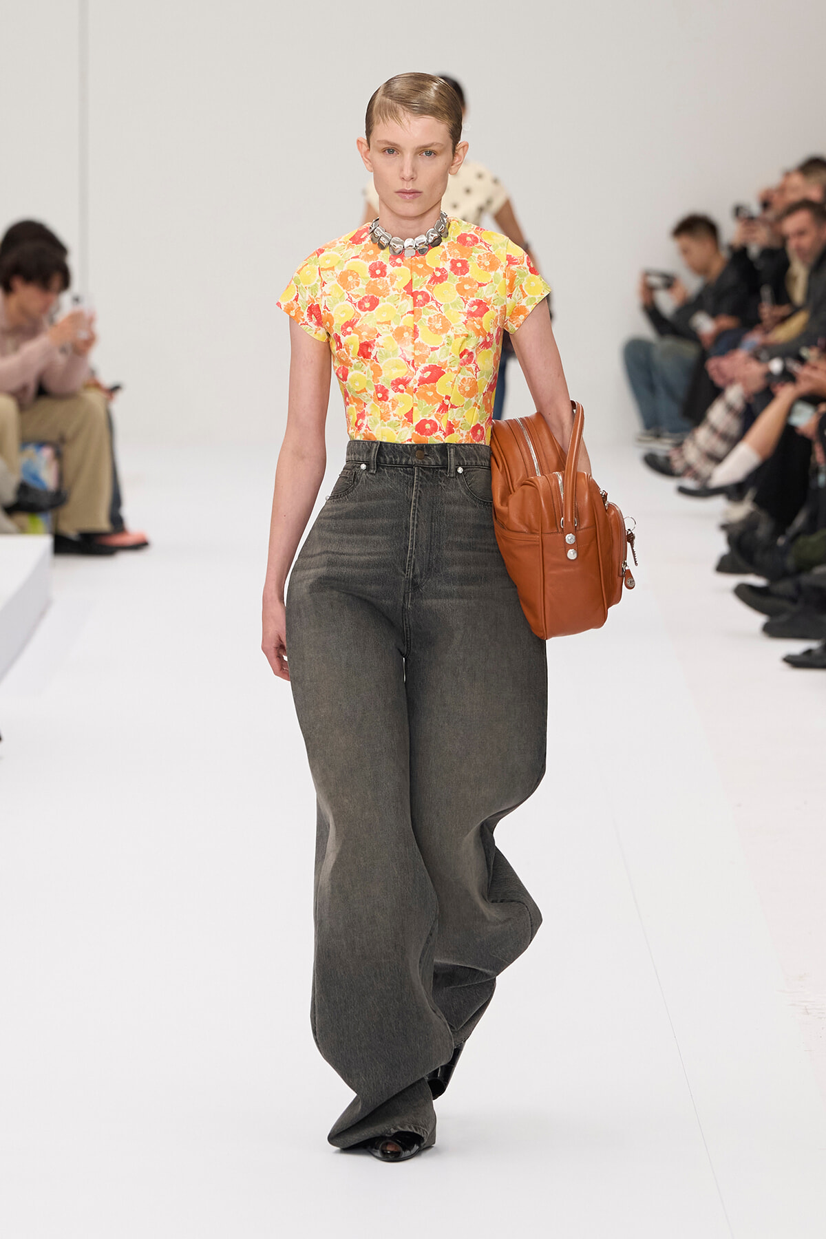 Model walks the runway in a yellow floral top and dark wide-leg jeans, carrying a brown backpack.
