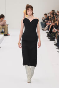 Model walks the runway in a strapless black midi dress with a heart-shaped neckline, paired with light gray knee-high boots.