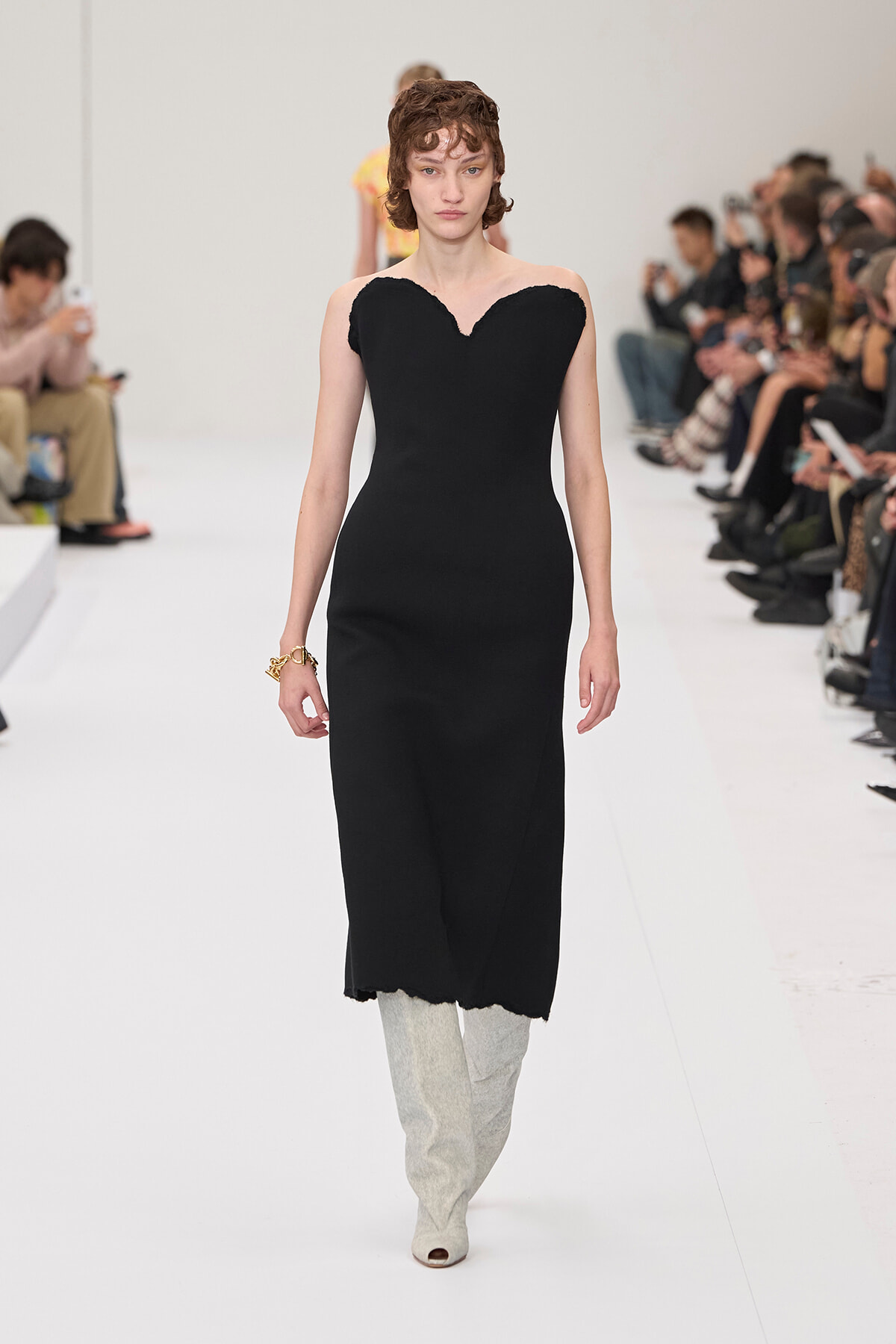 Model walks the runway in a strapless black midi dress with a heart-shaped neckline, paired with light gray knee-high boots.