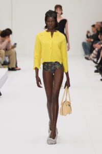 Model walking a fashion show wearing a bright yellow cardigan over floral bikini bottoms, glittery platform shoes, and carrying a small beige handbag.