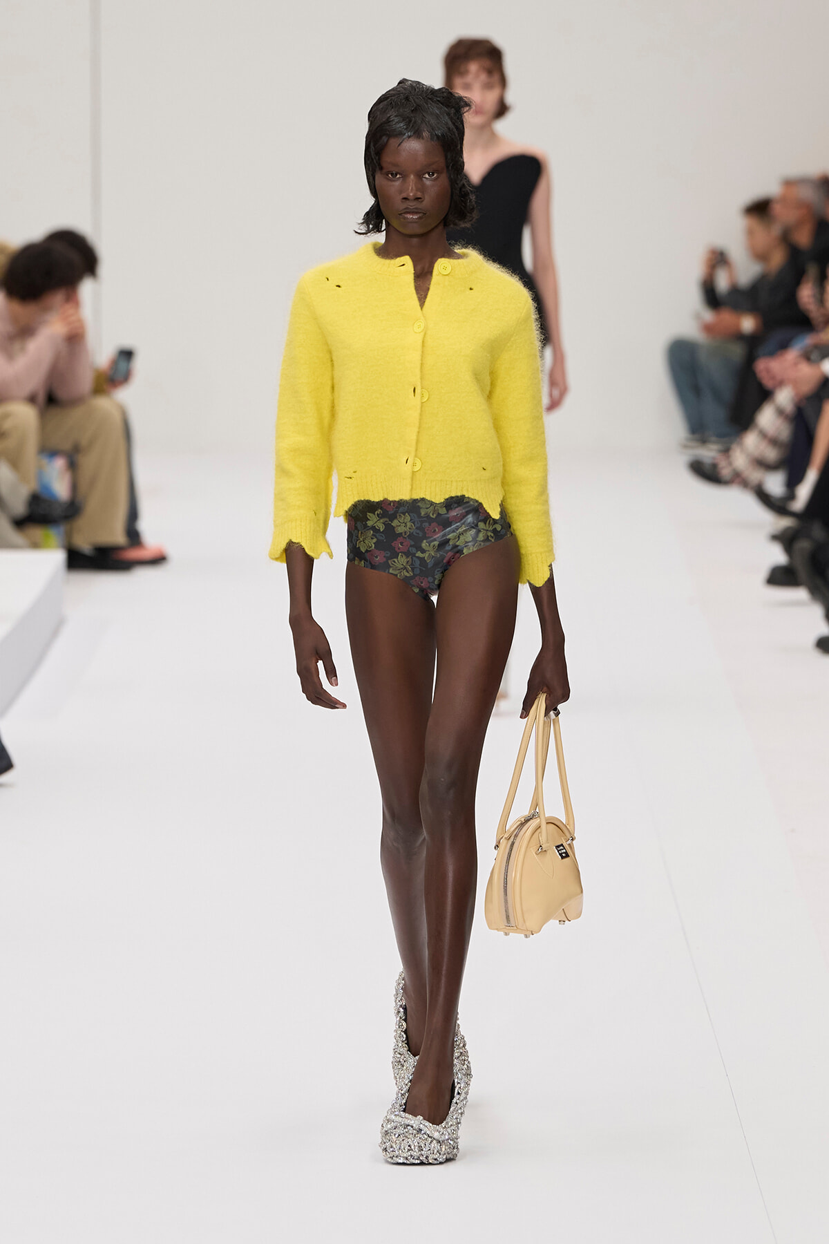 Model walking a fashion show wearing a bright yellow cardigan over floral bikini bottoms, glittery platform shoes, and carrying a small beige handbag.