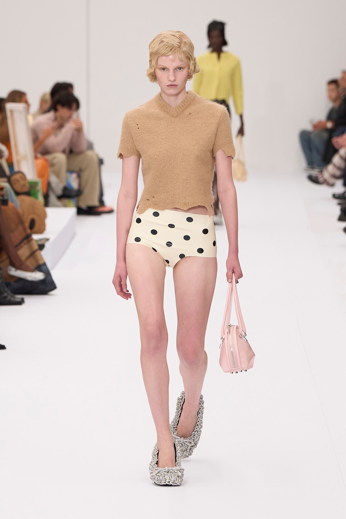 Model walking a fashion runway in a beige cropped knit top with scalloped sleeves and cream polka-dot high-waisted shorts, carrying a pink handbag.