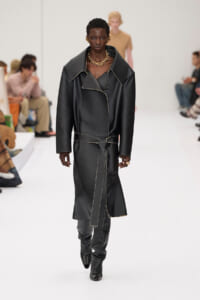 Model walks the runway in a black leather trench with a wrapped belt and gold jewelry.