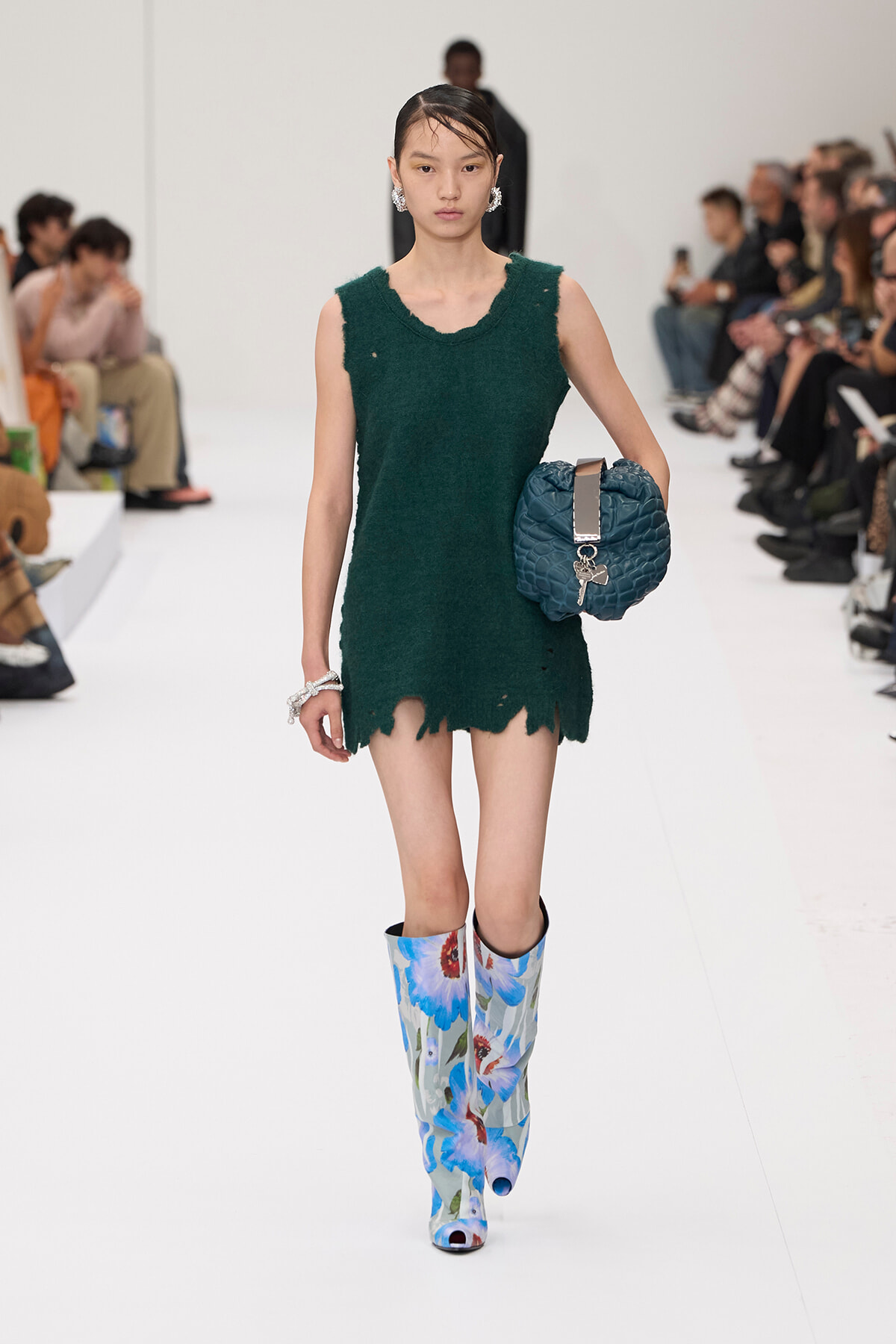Model walking a runway in a dark green distressed mini dress, blue floral knee-high boots, and a teal quilted bag with a metallic strap charm.