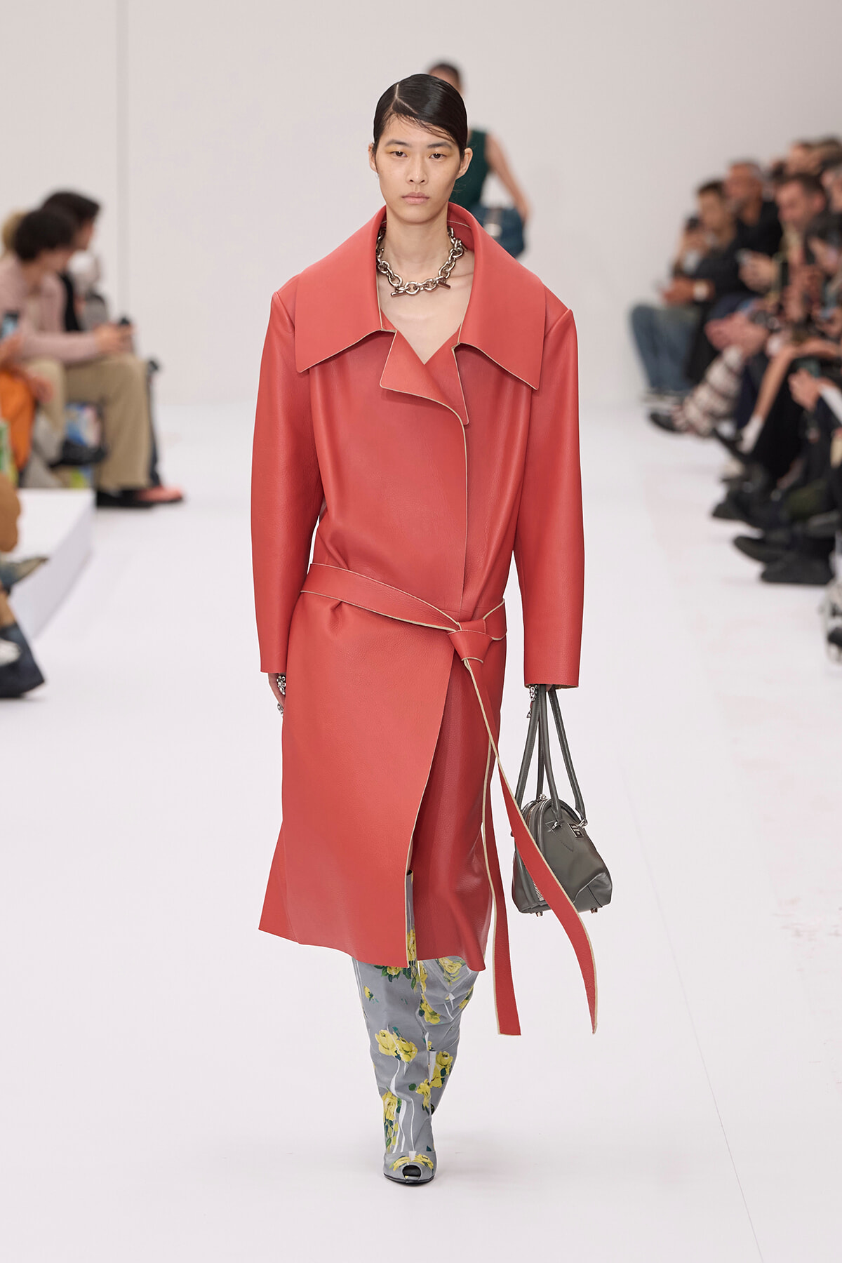 Model walks the runway in a coral pink oversized coat with a wide collar and belt, paired with floral grey pants and a grey handbag, audience visible in background.