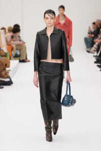 Model walking a fashion show in a black cropped jacket, beige top, and a matching black satin skirt, paired with floral boots and a blue handbag.