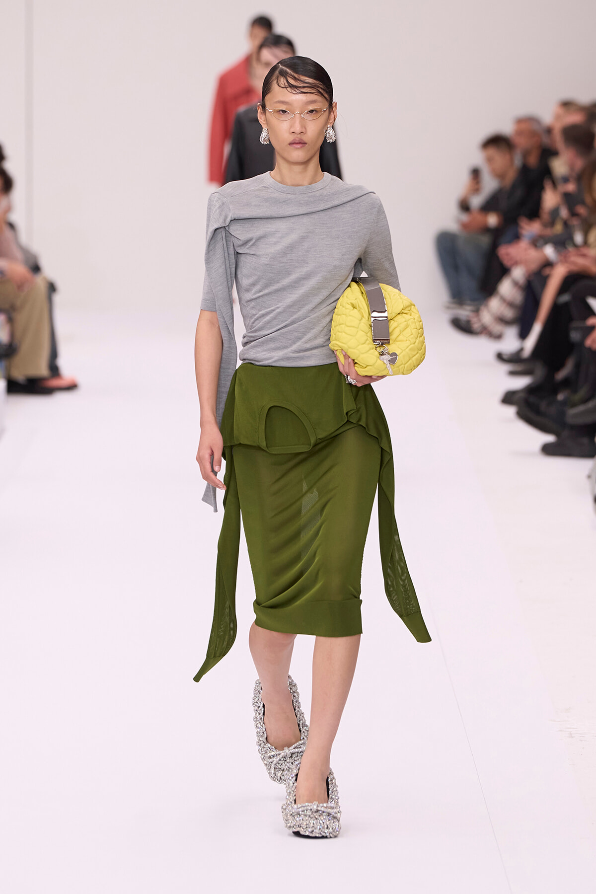 Model on a runway wearing a gray draped top and olive green layered midi skirt, holding a yellow quilted handbag with silver accessories and glittery heels.